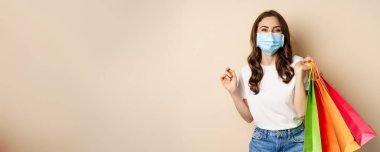 Covid-19 pandemic and lifestyle concept. Young woman posing in medical face mask with shopping bags from mall, vaccinated girl going in store in personal protective equipment, beige background.