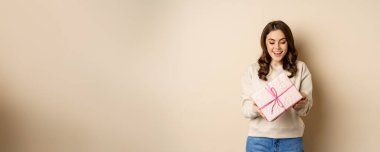 Excited beautiful girl with pink wrapped gift box, receive presents, standing over beige background.