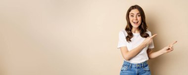 Smiling brunette woman in t-shirt, pointing fingers right, showing promo offer or advertisement, demonstrating banner, standing against beige background.