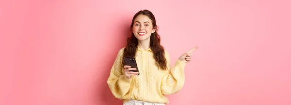 漂亮的现代女孩手握智能手机 把手指放在一边 一边用手机展示促销文字 一边站在粉色背景上 — 图库照片