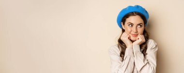 Close up portrait of feminine young woman in trendy french hat, posing cute and romantic, making silly coquettish face and tender smile, beige background.