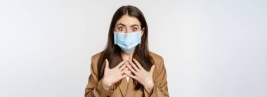 Workaplce ve pandemik konsept. Şok olmuş tıbbi maskeli iş kadını, soluk soluğa, kameraya korkmuş ve endişeli bakıyor, beyaz arka planda duruyor..