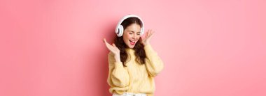 Mutlu kız en sevdiği şarkıyı söylüyor, kablosuz kulaklıkla müzik dinliyor, gülümsüyor ve dans ediyor, pembe arka planda duruyor..
