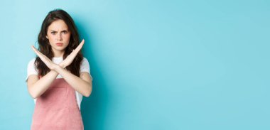 Hayal kırıklığına uğramış genç bir kadının surat asması, haç işareti yapması ve somurtması, dur hareketi göstermesi, hayır demesi, kötü davranışı yasaklaması, mavi arka planda durması..