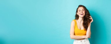 Yaz ve yaşam tarzı konsepti. Çekici bir kadının portresi gülüyor ve saçıyla oynuyor, sarı kesilmiş bluzla duruyor, tatile çıkıyor, mavi arka planda duruyor..