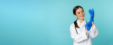 Smiling young woman, medical worker wearing gloves for patient checkup, standing in hospital clinic uniform over blue background.