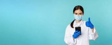 Online doctor and clinic. Young woman in medical face mask, using smartphone for client remote online appointment, showing thumbs up, standing over blue background.