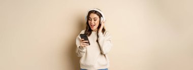 Smiling happy woman listening music in headphones on smartphone, laughing excited, standing over beige background.