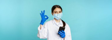 Online doctor and clinic. Young woman in medical face mask, using smartphone for client remote online appointment, showing okay sign, standing over blue background.