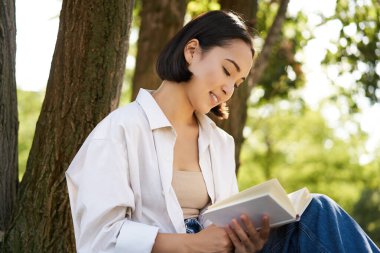 Güneşli bir günde parkta kitap okuyan güzel bir kız çimenlikte bir ağacın yanında oturup dışarıda dinleniyor..