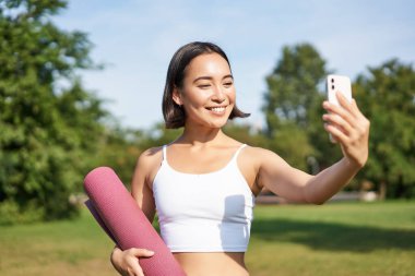 Sportif fitness kızı parkta lastik yoga minderiyle selfie çekiyor, spor yapıyor ve sosyal medyada fotoğraflarını paylaşıyor..