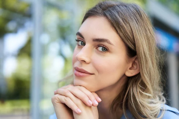 stock image Close up portrait of young woman, tuck hair behind ear, looking flirty and smiling, sitting in blue shirt outdoors on street.