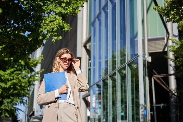 stock image Portrait of confident corporate woman going to office with a folder, walking on street on sunny day in suit and sunglasses.