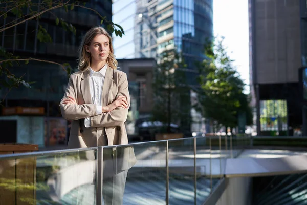 Confident businesswoman in suit, holds hands crossed on chest, stands in power pose on street.