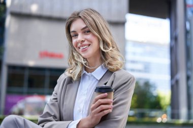 Meşgul bir caddede elinde kahveyle kameraya gülümseyen profesyonel bir kadın..