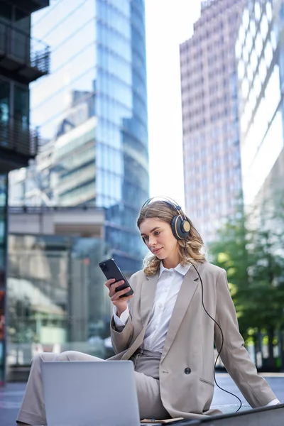 stock image Corporate woman in suit, sitting in city, listening to music with laptop, scrolling news on her mobile phone, relaxing on lunch break.