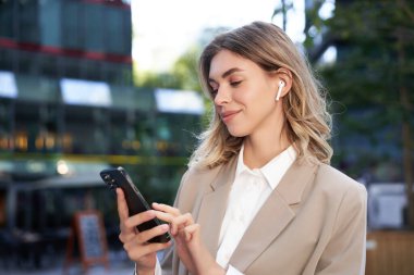 Bej takım elbise içinde gülümseyen güzel bir şirket kadını, şehirde kablosuz kulaklıklarla duruyor, akıllı telefon tutuyor..