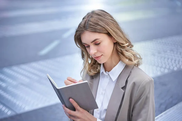 stock image Businesswoman in corporate suit, writes down smth, works on ideas in notebook, sits outside in city centre.