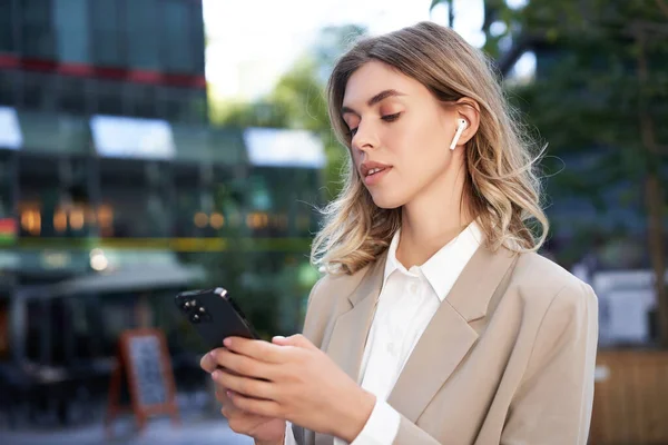 Stock image Successful businesswoman in beige suit, wireless headphones, looking at mobile phone, using smartphone app, standing outdoors on street.