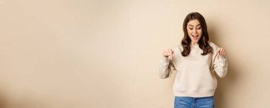 Excited adult woman pointing fingers down and smiling, looking at banner below, showing advertisement, found smth, standing over beige background.