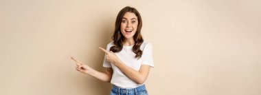 Smiling brunette woman in t-shirt, pointing fingers right, showing promo offer or advertisement, demonstrating banner, standing against beige background.