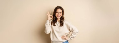 Excellent. Smiling beautiful woman showing okay, ok zero sign, approve smth good, praising and complimenting, beige background.