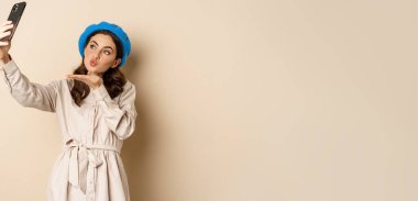Beautiful modern woman posing for selfie, holding smartphone, taking picture photo on mobile phone, standing happy against beige background.