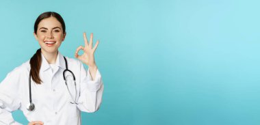 Portrait of satisfied, smiling medical worker, woman doctor showing okay, ok, zero no problem gesture, excellent sign, standing pleased over torquoise background.