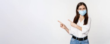 Tıbbi maskeli kurumsal bir kadının portresi, solu işaret ediyor, logoyu, şirket afişini gösteriyor, beyaz arka planda duruyor..