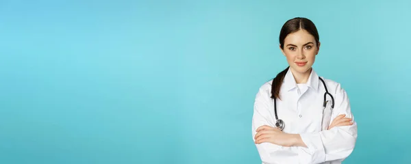 stock image Medical staff and doctors concept. Young smiling female doctor, healthcare worker in white coat and stethoscope, looking confident, waiting patients, blue background.