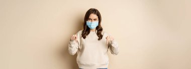 Angry woman in medical face mask, pointing fingers down and looking concerned, standing in sweater over beige background.