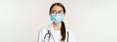 Portrait of medical worker, female physician in face mask from covid during pandemic, smiling and looking enthusiastic, standing over white background.
