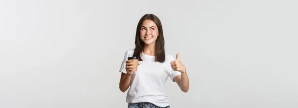 迷人的面带微笑的黑发女孩看上去很满意 喝咖啡 竖起大拇指 推荐咖啡馆 — 图库照片