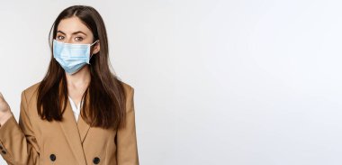 Coronavirus and people concept. Portrait of business woman at workplace wearing face mask, pointing finger left at logo, company banner, white background.