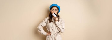 Shopping and discounts concept. Young stylish modern girl, posing with credit card, looking happy and smiling, standing over beige background.