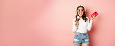Yazlık kız gülüyor, lolipop yiyor ve kulaklıkla müzik dinliyor, pembe arka planda akıllı telefonla dans ediyor..