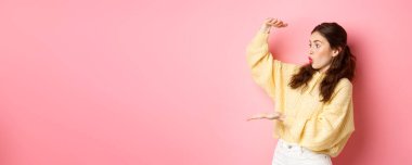 Esmer kızın portresi büyük bir boşluk tutuyor ve ürününüz veya logonuz için hayret verici bir yere bakıyor, büyük bir kutu hareketi gösteriyor, pembe arka planda duruyor..