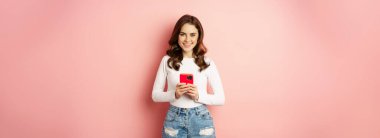 Online shopping and cellphone concept. Excited attractive girl holding mobile phone and smiling, standing over pink background.