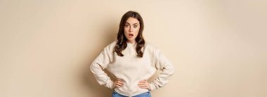 Portrait of shocked brunette woman drop jaw, gasping and staring speechless at camera, beige background. Copy space