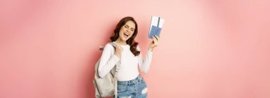 Vacation and holiday. Beautiful girl going on trip, holding passport with airplane tickets, holding backpack, travelling, standing over pink background.