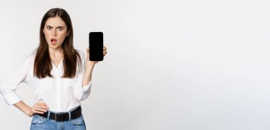 Shocked and frustrated woman showing mobile phone screen app, smartphone application, looking insulted, standing over white background.
