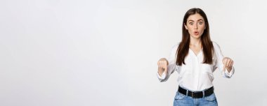 Surprsed businesswoman, beautiful adult woman pointing fingers down, looking excited at camera, standing over white background.