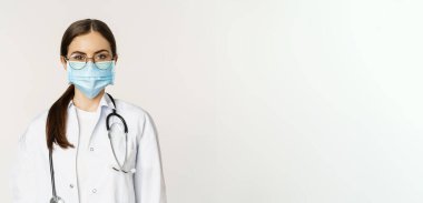 Kadın doktor portresi, gözlüklü hastane personeli ve tıbbi maske, ciddi görünüyor, hastaları karşılamaya hazır, beyaz arka planda duruyor..