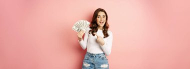 Attractive young woman holding money, cash in hands, concept of loans, microcredit and shopping, standing over pink background. Copy space