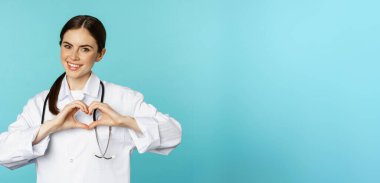 Young doctor, woman physician showing heart, love sign and smiling, care for patients in clinic, standing in white coat over blue background.