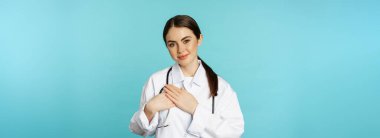 Caring healthcare worker, young female doctor holding hands on heart and smiling, looking with care and tenderness, standing over torquoise background.