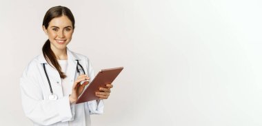 Young professional doctor, woman physician with stethoscope, holding digital tablet and smiling at camera, working in clinic, standing over white background.