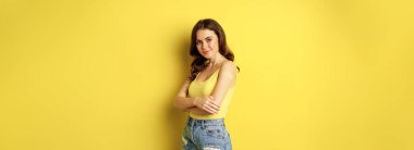 Vaccination campaign concept. Cute smiling young woman after getting vaccinated, standing with patch on her shoulder and looking healthy and happy, yellow background.