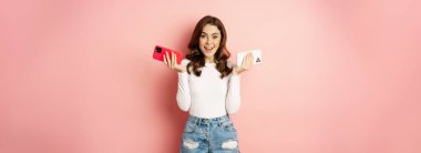 Technology, spring promo concept. Stylish glamour woman holding two smartphones, mobile phones in both hands, smiling pleased, buying new cellphone, pink background.
