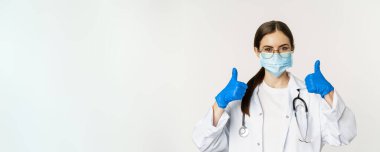 Coronavirus 'un yüz maskesi takmış kadın doktorların portresi. Baş parmaklarını gösterip gülümsüyor. Klinik ya da tıbbi web sitesi tavsiye ediyor..